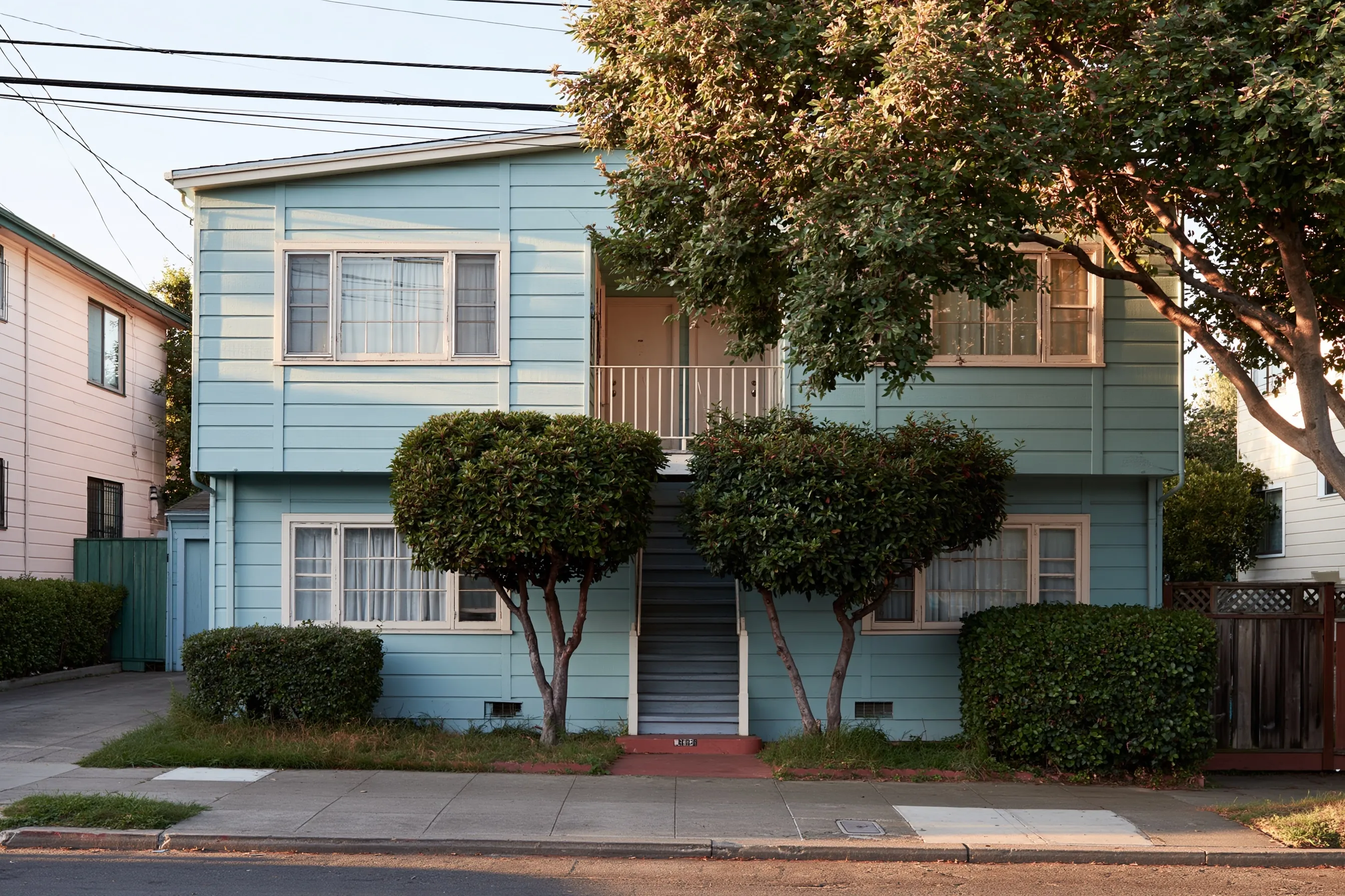 Two-story apartment building with painted wood siding, wood-framed windows and an exterior stair, two trimmed shrubs in front